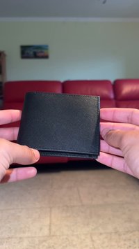 Portafoglio in pelle nera con superficie texturizzata, design a due pieghe e cuciture visibili lungo i bordi, tenuto tra due mani.