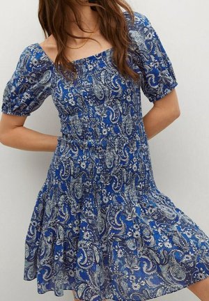 Femme portant une robe bleue avec des motifs floraux et paisley blancs, des manches courtes bouffantes et un décolleté carré, debout devant un fond uni.