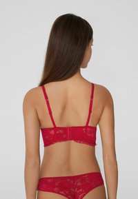 Červená krajková podprsenka a odpovídající tanga, vybavené nastavitelnými ramínky, uzavřením na háček a očko a detailem s vlnitým okrajem. Hladký materiál.