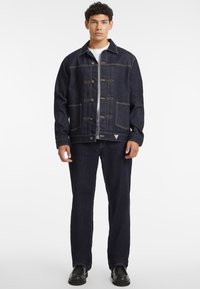 Giacca di denim scuro con un design strutturato, cuciture a contrasto, grandi tasche frontali e chiusura a bottoni. Abbinata a pantaloni di denim coordinati.