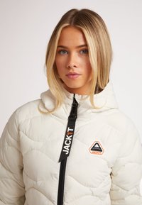 Vit quiltad puffjacka med huva, som har dragkedja framtill, svart rem med texten "JACKIT" och en diamanlogotyp.