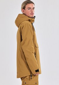 Brown waterdichte jas met een capuchon, voorzakken en verstelbare trekkoorden. Gladde textuur en strak ontwerp, geschikt voor buitengebruik.