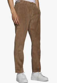 Brune fløjlsbukser med vertikal ribbet tekstur, elastisk talje, snøre og sidelommer, parret med hvide sneakers.