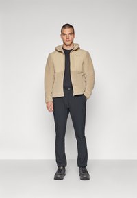 Beige Fleece-Jacke mit einem kontrastierenden glatten Stoff an der Vorderseite, Reißverschluss und Kapuze. Kombiniert mit schwarzen Hosen und dunklen Schuhen.