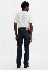Jeans de mezclilla azul oscuro con pierna recta, parche de cuero marrón en la cintura trasera y detalles de costura amarilla. Combinados con una camiseta blanca.