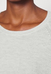 Hellgrauer, gestrickter Pullover mit weitem Rundhalsausschnitt, der ein strukturiertes, offenes Gewebe-Muster zeigt. Glattes Material und lockere Passform.