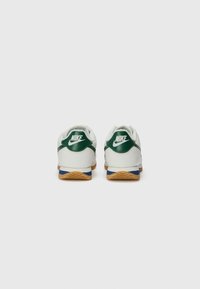 Ténis Nike brancos com detalhes em verde, apresentando um calcanhar em pele texturada, detalhes azuis na sola e uma sola em borracha de alperce.