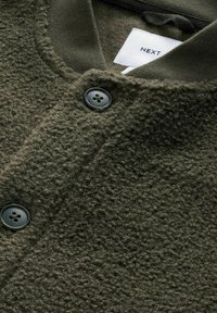 Olive green textured fleece jacket with a smooth collar, featuring two large black buttons and a tag labeled 'NEXT'.