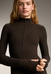 Žena s dugom plavom kosom, odjevena u uski tamnosmeđi rebrasti džemper sa prednjim patentnim zatvaračem i otvorima za palce, držeći jedan rukav.