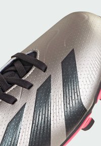 Botas de fútbol con una parte superior metálica plateada que presenta acentos de rayas negras y superficies texturizadas, cordones negros y una suela rosa.