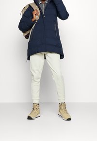 Granatowa kurtka puchowa z kapturem, białe spodnie i beżowe buty. Plecak jest jasnozłoty, z regulowanymi paseczkami i zamkami błyskawicznymi.