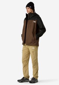 Brown and black waterproof jacket with a hood, beige pants, and black shoes. The jacket features logo accents and zippered pockets.