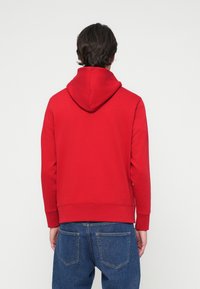 Sweatshirt vermelho com capuz, de textura suave, com punhos e bainha canelados. Estilizado com jeans azuis, visto de costas contra um fundo neutro.