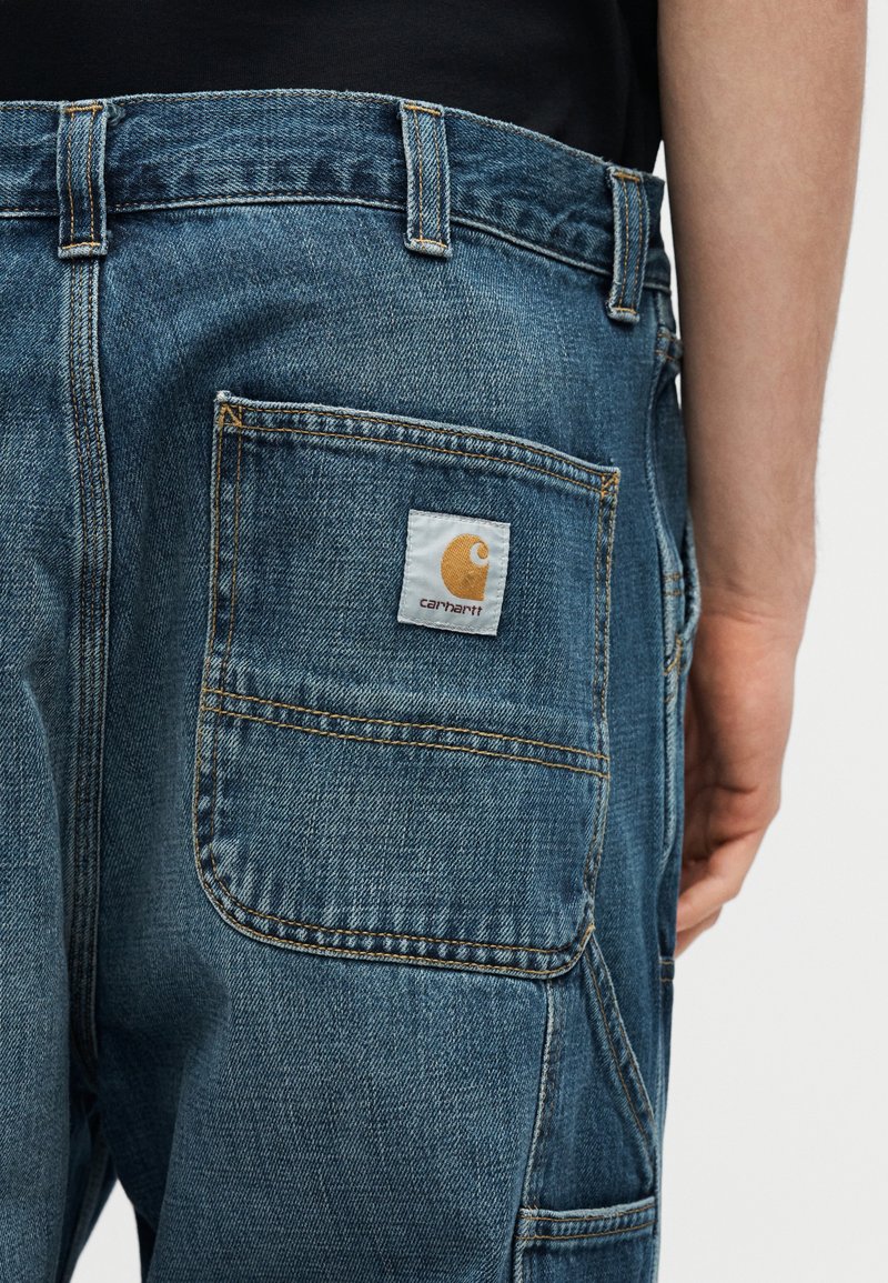 Pantalones vaqueros de mezclilla azul con un bolsillo trasero que tiene una etiqueta blanca con el logo amarillo de Carhartt, usados con una camisa negra visible en la cintura.