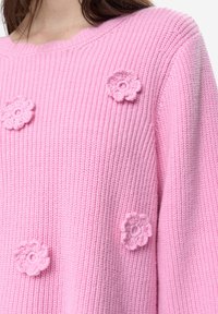 Růžový pletený svetr s texturovaným vzorem, kulatým výstřihem a trojrozměrnými květinovými ozdobami ve stejném odstínu.
