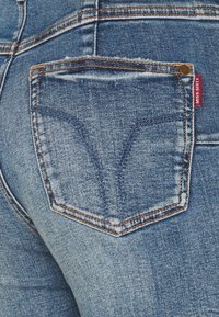 Jeans de mezclilla azul con un diseño distintivo en el bolsillo trasero, que presenta costuras naranjas y una pequeña etiqueta roja en el lateral.