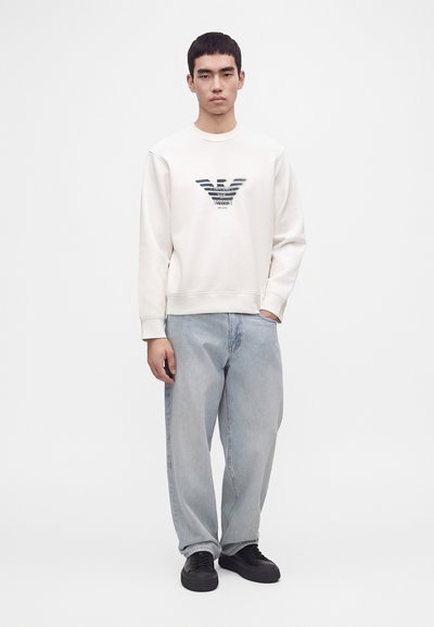 Jeune homme debout portant un sweat-shirt blanc avec un logo noir, un jean large bleu clair et des baskets noires sur un fond uni clair.