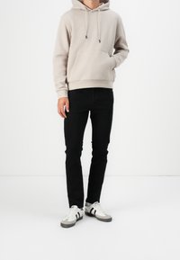 Felpa beige con tasca frontale e cappuccio regolabile, abbinata a jeans neri slim-fit e sneakers bianche con strisce grigie e nere.