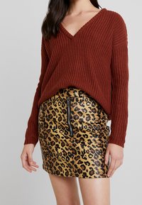 Vrouw in een roestkleurige chunky gebreide V-hals trui, gestopt in een glanzende mini-jurk met luipaardprint, met een rits aan de voorkant.