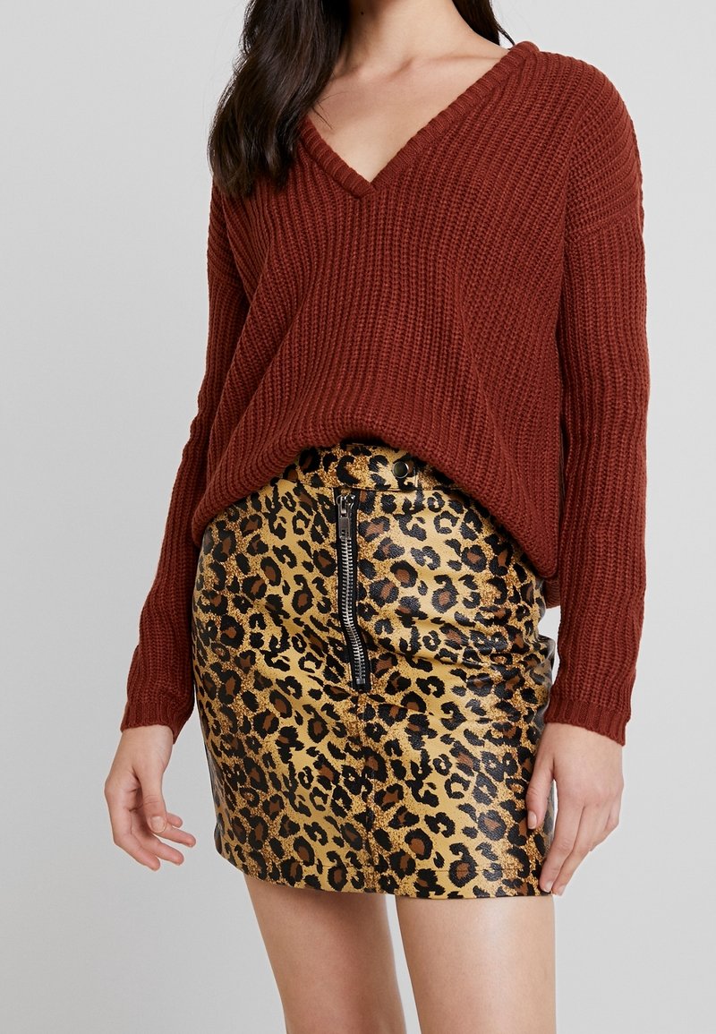Vrouw in een roestkleurige chunky gebreide V-hals trui, gestopt in een glanzende mini-jurk met luipaardprint, met een rits aan de voorkant.