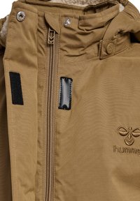 Brun jakke med glidelås, trykknapper og borrelåsdetaljer; myk, teksturert fôr; brodert logo på forsiden.