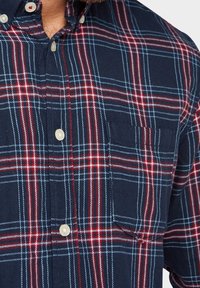 Camicia a quadri navy con motivi a scacchi rossi e bianchi, chiusura con bottoni e un'unica tasca sul petto. Tessuto morbido con finitura testurizzata.