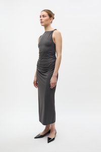 Femeie în picioare purtând o rochie midi gri închis fără mâneci, cu detalii strânse și guler înalt, combinată cu pantofi negri cu vârf ascuțit, pe fundal alb.