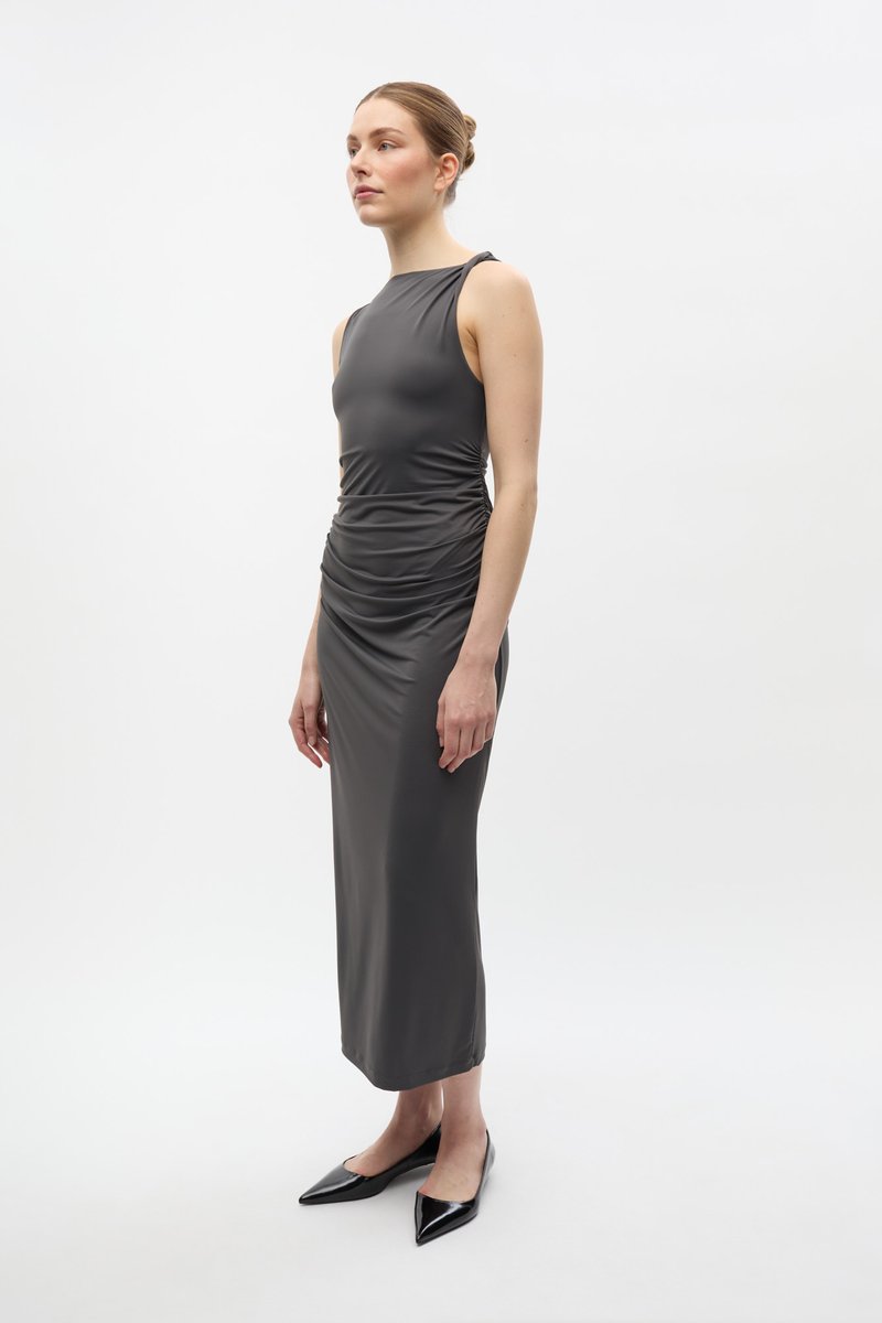 Femeie în picioare purtând o rochie midi gri închis fără mâneci, cu detalii strânse și guler înalt, combinată cu pantofi negri cu vârf ascuțit, pe fundal alb.