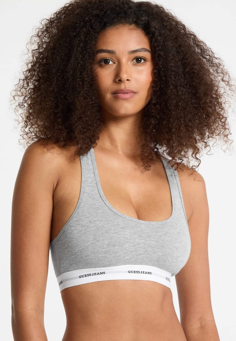 Grå bomullssportbehå med racerback-design och ett vitt elastiskt band med texten "GUESS JEANS" i svart. Slät textur och åtsittande form.