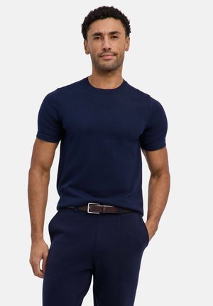 Mann mit kurzen lockigen Haaren, der ein eng anliegendes, marineblaues Kurzarmhemd und passende Hosen trägt und mit einer Hand in der Tasche steht.