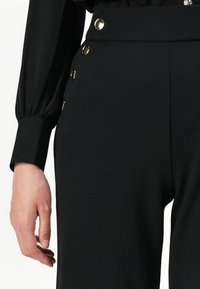 Femme portant un pantalon noir taille haute avec des boutons dorés sur le côté et une blouse noire à manches longues avec de larges poignets.