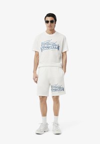 Conjunto de camiseta de algodón blanca y pantalones cortos que presenta un gráfico del logo azul de Lacoste. Llevado con gafas de sol y calzado blanco.