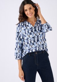 Camicia button-up azzurra chiara con motivi geometrici astratti in navy e bianco, caratterizzata da maniche a tre quarti e una vestibilità rilassata. Abbinata a jeans scuri.