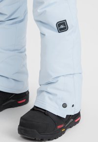 Gaiši zili sniega bikses, kas izgatavotas no ūdensnecaurlaidīga materiāla, ar melnu O’Neill logotipu un pogu detaļu pie potītes, saskaņotas ar melnām zābakām.
