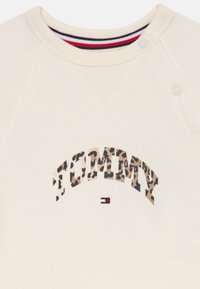 Grietos spalvos džemperio apykaklė su užsegamais mygtukais, leopardų rašto tekstas "TOMMY" ir nedidelis raudonas, baltas ir mėlynas vėliavos logotipas žemiau.