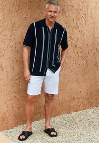 Camisa de manga corta de la marina con rayas verticales blancas y detalles texturizados, combinada con pantalones cortos blancos y sandalias negras sin hebilla.