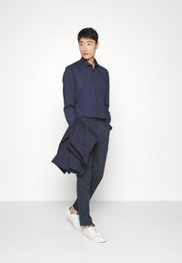 Mørkeblå rutete knapp-skjorte kombinert med mørkeblå bukser, en mørkeblå blazer drapert over armen, og hvite sneakers med grønne detaljer.