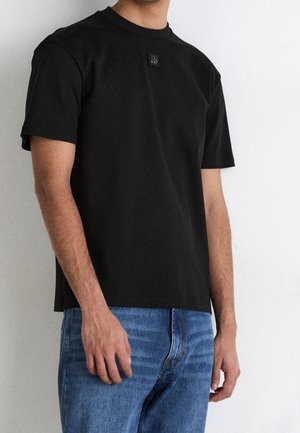 Czarny T-shirt z krótkim rękawem wykonany z bawełny, z okrągłym dekoltem i prostokątną naszywką z logo na górnej części klatki piersiowej. Założony z niebieskimi dżinsami.