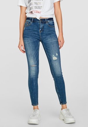 Mujer con vaqueros ajustados azules con parches desgastados, una camiseta gráfica blanca anudada y zapatillas deportivas blancas gruesas, de pie contra un fondo liso.