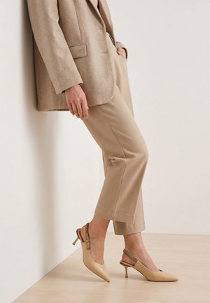 Kvinde iført beige skræddersyede bukser, matchende blazer og beige slingback kitten heels, stående mod en hvid væg på en lyst trægulv.