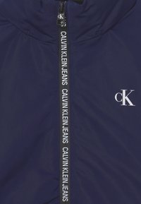 Námořnické modré prošívané sako s vysokým límcem, černým zipem a bílým logem. Obsahuje vertikální bílou logovou pásku podél zipu.