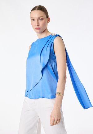 Femme portant un chemisier sans manches bleu vif avec un panneau arrière fluide et un bracelet doré, assorti à un pantalon blanc sur un fond uni.