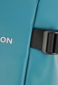 Matériau imperméable turquoise avec une finition texturée et une boucle réglable noire. Le nom de la marque est embossé en blanc sur la surface.