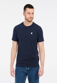 Tmavomodré bavlnené tričko s okrúhlym výstrihom, krátkymi rukávmi a náprsné vrecko s malou nášivkou. Nosené s svetlomodrými džínami.