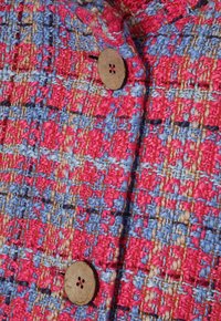 Un tissu tissé multicolore présente des fils roses et bleus vifs. Un bouton en bois rond est visible, accentuant le motif texturé.