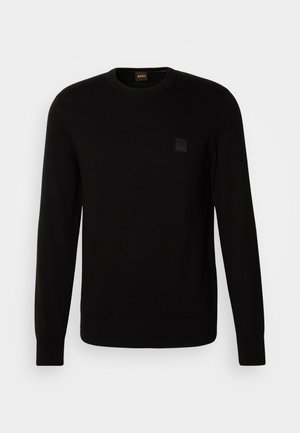 Suéter negro de punto con mangas largas, escote redondo y un pequeño parche de logo texturizado en el pecho. Textura suave en todo.