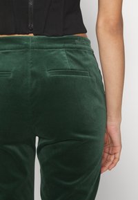 Zelené sametové kalhoty s kapsami na zadní straně; hladká textura a přizpůsobený střih. Nosit s černým zkráceným topem.