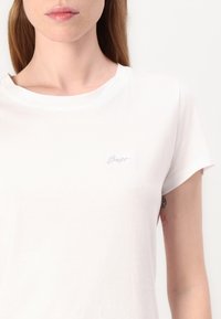 Tricou alb din bumbac cu guler rotund, mâneci scurte și un mic logo brodat pe piept. Textură netedă, design casual.