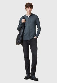 Felpa grigia con zip e lacci, indossata sopra una camicia bianca. Abbinata a jeans neri e scarpe nere. Il modello tiene in mano una borsa nera.