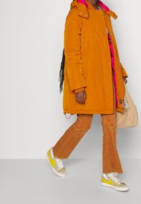 Personne marchant en pantalon en velours côtelé orange brûlé et manteau assorti, portant des baskets jaunes vives et blanches, tenant un sac à main beige pelucheux.